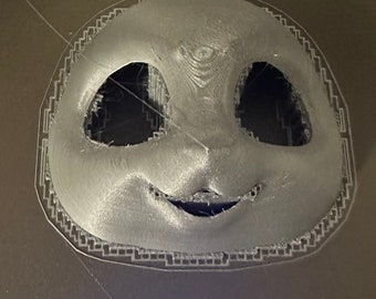 May include: A grey 3D-printed mask with large eye openings and a smiling mouth. The mask has a rounded shape with a decorative border. The surface has visible print lines, indicating a 3D-printing process.