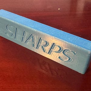 Op de afbeelding: Een rechthoekig, lichtblauw object met het woord "SHARPS" in de bovenkant gegraveerd. Het object ligt op een donkerrood oppervlak.