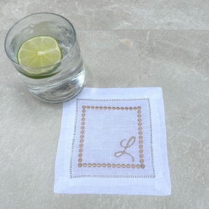May include: White linen cocktail napkin with a brown embroidered border and the letter "L" in the center.