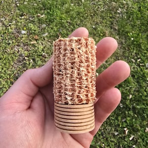 Può includere: Oggetto cilindrico con una sezione superiore strutturata e intrecciata e una base composta da pezzi di legno circolari impilati. L'oggetto è tenuto in mano su uno sfondo di erba verde.