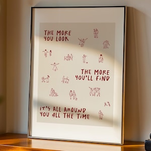 Op de afbeelding: Ingelijste kunstdruk met de tekst "THE MORE YOU LOOK, THE MORE YOU'LL FIND, IT'S ALL AROUND YOU ALL THE TIME" in het rood. De print bevat kleine rode illustraties van mensen. De lijst is zwart en staat op een houten plank.