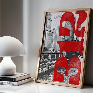 May include: A framed art print featuring a black and white cityscape with bold red text overlay. The artwork is displayed on a white surface, next to a white mushroom-shaped lamp and two books. The frame is made of light-colored wood.