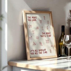 Puede incluir: Una impresi&oacute;n art&iacute;stica enmarcada con el texto "THE MORE YOU LOOK, THE MORE YOU'LL FIND, IT'S ALL AROUND YOU ALL THE TIME" en rojo. La impresi&oacute;n est&aacute; en un marco de madera clara y se exhibe en una encimera junto a una botella y una copa de vino.