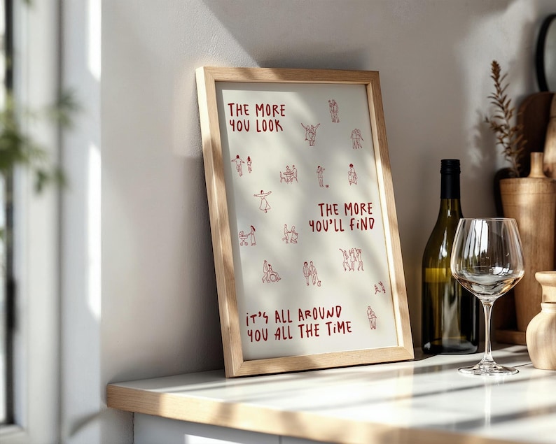 Puede incluir: Una impresi&oacute;n art&iacute;stica enmarcada con el texto "THE MORE YOU LOOK, THE MORE YOU'LL FIND, IT'S ALL AROUND YOU ALL THE TIME" en rojo. La impresi&oacute;n est&aacute; en un marco de madera clara y se exhibe en una encimera junto a una botella y una copa de vino.