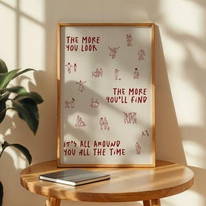 Puede incluir: Un marco de madera que contiene una impresi&oacute;n con un fondo claro. La impresi&oacute;n presenta dibujos lineales rojos de personas y el texto "THE MORE YOU LOOK, THE MORE YOU'LL FIND, IT'S ALL AROUND YOU ALL THE TIME." Un cuaderno cerrado descansa sobre una mesa de madera.