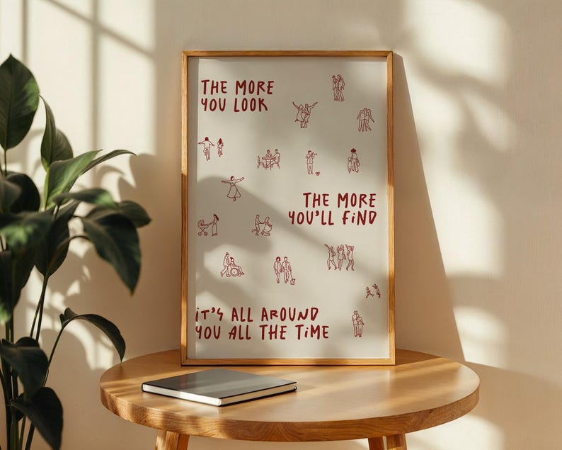 Puede incluir: Un marco de madera que contiene una impresi&oacute;n con un fondo claro. La impresi&oacute;n presenta dibujos lineales rojos de personas y el texto "THE MORE YOU LOOK, THE MORE YOU'LL FIND, IT'S ALL AROUND YOU ALL THE TIME." Un cuaderno cerrado descansa sobre una mesa de madera.