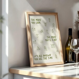 May include: A framed print with the text "THE MORE YOU LOOK, THE MORE YOU'LL FIND, IT'S ALL AROUND YOU ALL THE TIME" in green. The print is in a light wood frame and is displayed on a countertop next to a wine bottle and a wine glass.