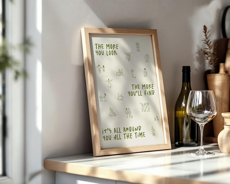 May include: A framed print with the text "THE MORE YOU LOOK, THE MORE YOU'LL FIND, IT'S ALL AROUND YOU ALL THE TIME" in green. The print is in a light wood frame and is displayed on a countertop next to a wine bottle and a wine glass.