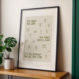 May include: Framed art print with a black frame. The print has a beige background with the text "THE MORE YOU LOOK THE MORE YOU'LL FIND IT'S ALL AROUND YOU ALL THE TIME" in green. Small green line drawings of people are scattered across the print.