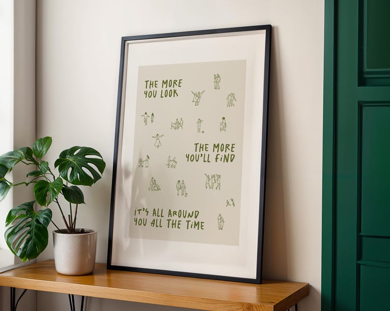 May include: Framed art print with a black frame. The print has a beige background with the text "THE MORE YOU LOOK THE MORE YOU'LL FIND IT'S ALL AROUND YOU ALL THE TIME" in green. Small green line drawings of people are scattered across the print.