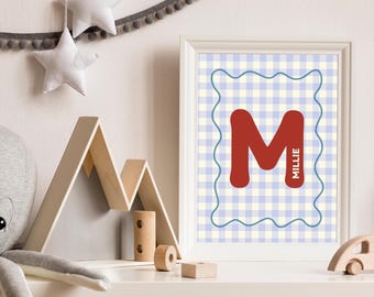 May include: A framed print with a red letter 'M' and the name 'Millie' in white, set against a blue and white gingham background. The print is in a white frame. Wooden toys and star decorations are also visible in the image.