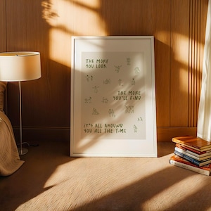 May include: A framed art print with the text "THE MORE YOU LOOK, THE MORE YOU'LL FIND, IT'S ALL AROUND YOU ALL THE TIME" in green. The print is displayed in a room with a lamp, bed, and a stack of books.