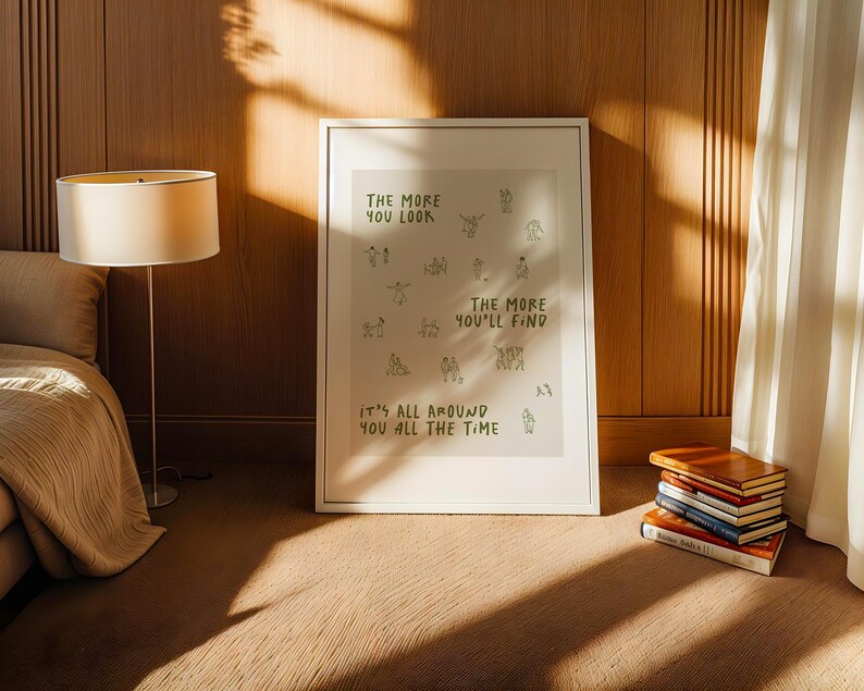 May include: A framed art print with the text "THE MORE YOU LOOK, THE MORE YOU'LL FIND, IT'S ALL AROUND YOU ALL THE TIME" in green. The print is displayed in a room with a lamp, bed, and a stack of books.