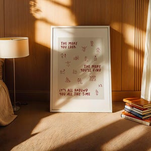 Puede incluir: Un cuadro enmarcado con el texto rojo "THE MORE YOU LOOK, THE MORE YOU'LL FIND, IT'S ALL AROUND YOU ALL THE TIME". El cuadro est&aacute; en una habitaci&oacute;n con una cama, una l&aacute;mpara y una pila de libros.