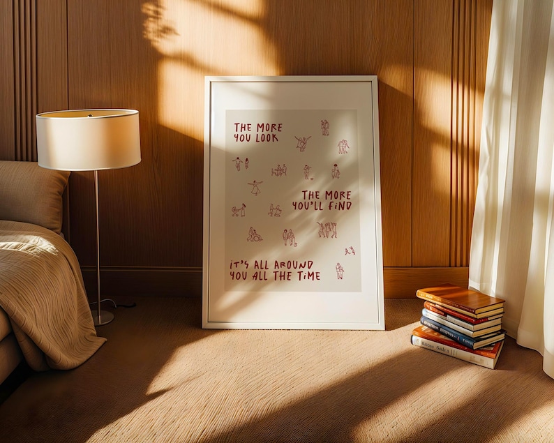 Puede incluir: Un cuadro enmarcado con el texto rojo "THE MORE YOU LOOK, THE MORE YOU'LL FIND, IT'S ALL AROUND YOU ALL THE TIME". El cuadro est&aacute; en una habitaci&oacute;n con una cama, una l&aacute;mpara y una pila de libros.