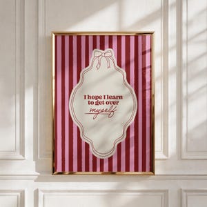 May include: A rectangular wall art print in a gold frame featuring a vertical striped pattern in shades of pink and dark red. Centered is a cream-colored scalloped label shape with a bow at the top, containing the text I hope I learn to get over myself.