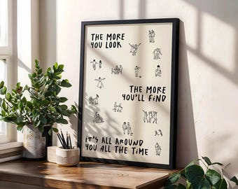 May include: A framed art print with a black border, featuring the text "THE MORE YOU LOOK, THE MORE YOU'LL FIND, IT'S ALL AROUND YOU ALL THE TIME." The print has simple black line drawings of people. The print is on a wooden surface.