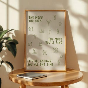 May include: A framed art print with a light background and a wooden frame. The print features green text and line drawings of people. The text reads "THE MORE YOU LOOK, THE MORE YOU'LL FIND, IT'S ALL AROUND YOU ALL THE TIME."