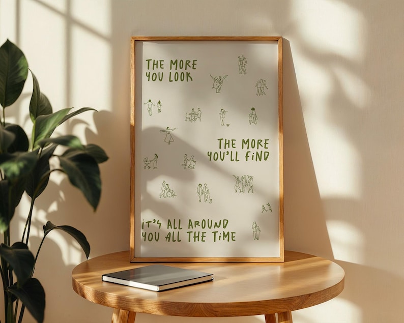 May include: A framed art print with a light background and a wooden frame. The print features green text and line drawings of people. The text reads "THE MORE YOU LOOK, THE MORE YOU'LL FIND, IT'S ALL AROUND YOU ALL THE TIME."