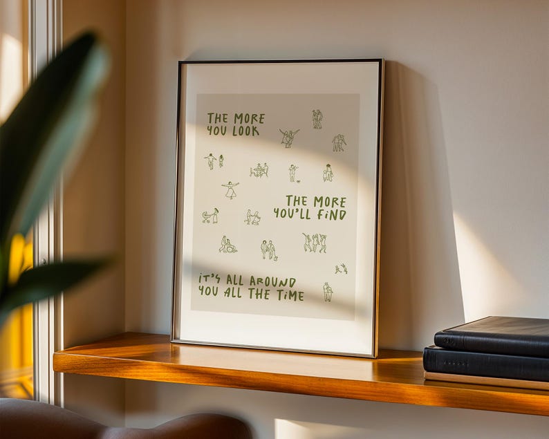 May include: A framed art print with a white background and black text that reads "THE MORE YOU LOOK THE MORE YOU'LL FIND IT'S ALL AROUND YOU ALL THE TIME." The print has small green illustrations of people. The frame is black.