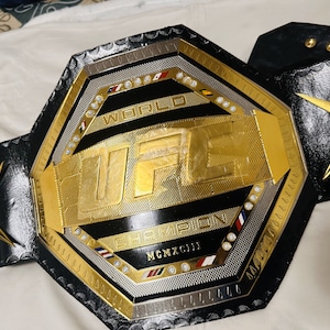 May include: A gold and black UFC World Champion belt with a black leather strap and silver accents. The belt features the UFC logo and the words "World Champion" and "MCMXCIII".