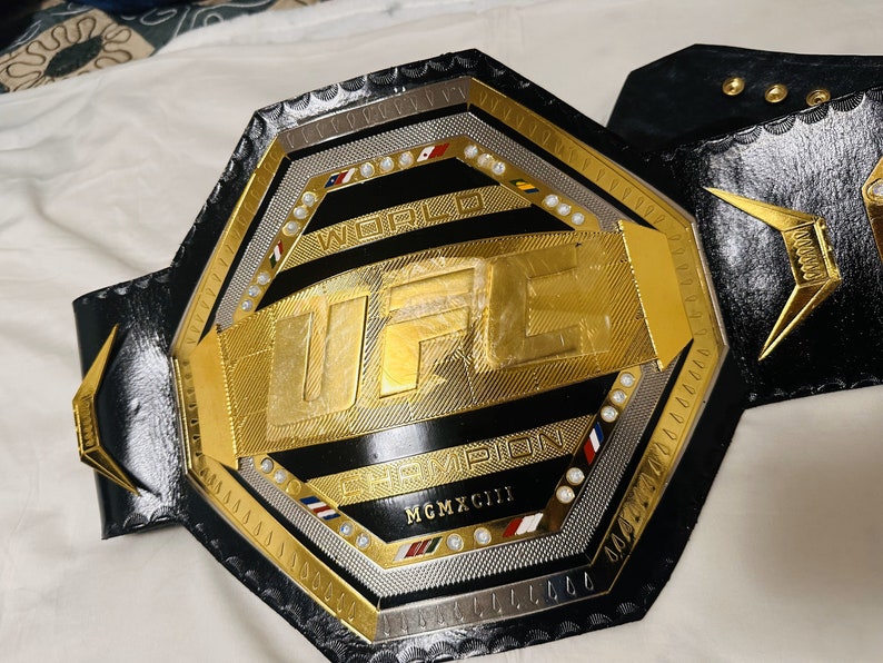May include: A gold and black UFC World Champion belt with a black leather strap and silver accents. The belt features the UFC logo and the words "World Champion" and "MCMXCIII".