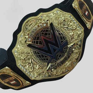 May include: A gold and black wrestling championship belt with a large, ornate center plate featuring a black "W" logo and the words "World Heavyweight Champion". The belt has a black leather strap with gold accents and two side plates with the word "GUNTHER" on them.