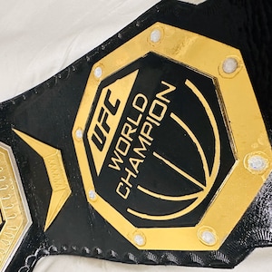 May include: A gold and black UFC World Champion belt with a black octagon-shaped plate featuring the UFC logo and the words "World Champion".