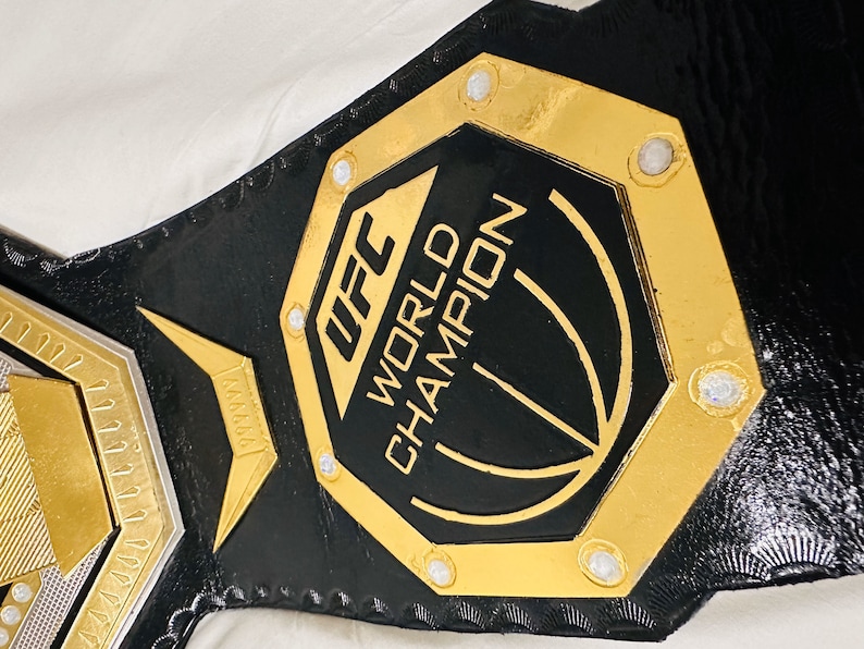 May include: A gold and black UFC World Champion belt with a black octagon-shaped plate featuring the UFC logo and the words "World Champion".