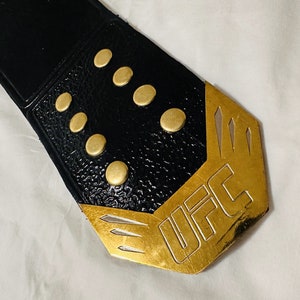 May include: Close-up of a black leather UFC championship belt with gold accents and the word "UFC" engraved on the buckle.