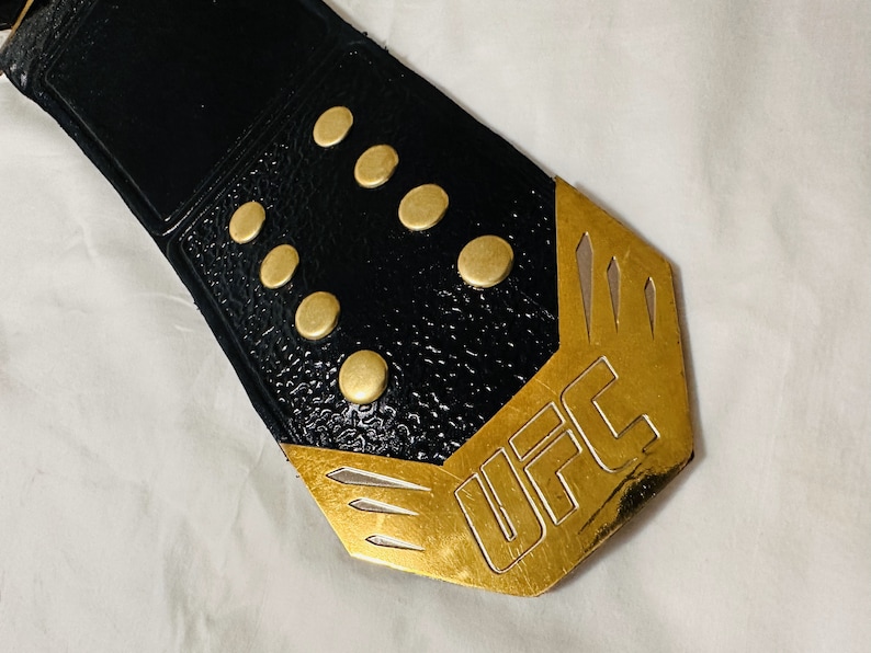 May include: Close-up of a black leather UFC championship belt with gold accents and the word "UFC" engraved on the buckle.
