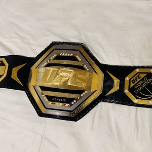 May include: A black leather UFC World Champion belt with a gold octagon-shaped buckle. The buckle features the UFC logo, the words "World Champion", and the year "MCMXCIII".