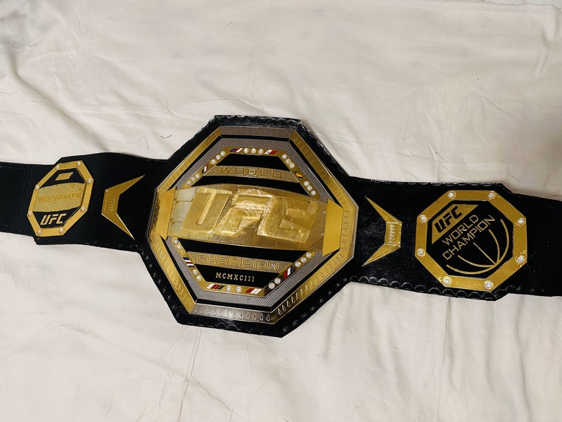 May include: A black leather UFC World Champion belt with a gold octagon-shaped buckle. The buckle features the UFC logo, the words "World Champion", and the year "MCMXCIII".