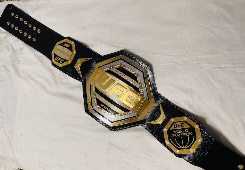 May include: A black leather UFC World Champion belt with a gold octagon-shaped buckle. The buckle features the UFC logo and the words "World Champion".
