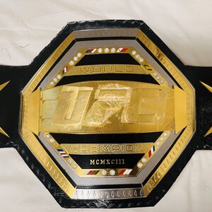 May include: A gold UFC championship belt with black leather straps and a silver octagon-shaped center plate. The center plate features the UFC logo, the words "World Champion", and the year "MCMXCIII".