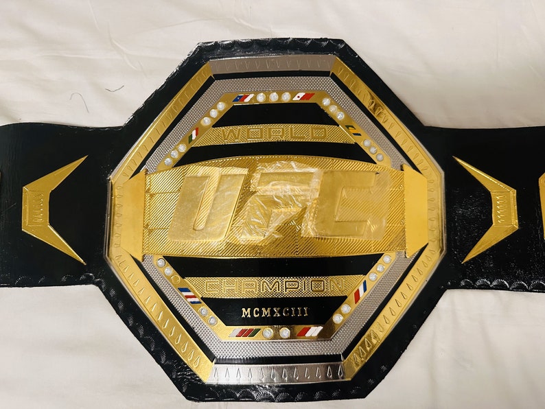 May include: A gold UFC championship belt with black leather straps and a silver octagon-shaped center plate. The center plate features the UFC logo, the words "World Champion", and the year "MCMXCIII".