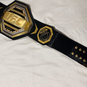 May include: A black leather UFC championship belt with gold accents. The belt features a large octagon-shaped buckle with the UFC logo and the words "World Champion".
