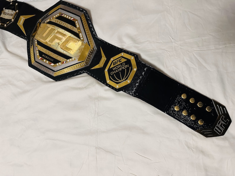 May include: A black leather UFC championship belt with gold accents. The belt features a large octagon-shaped buckle with the UFC logo and the words "World Champion".
