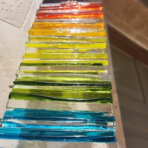 May include: An abstract glass artwork with a gradient of colours. The piece is composed of horizontal glass strips, transitioning from red to orange, yellow, green, and blue. The glass has a reflective quality, creating a vibrant and textured appearance.