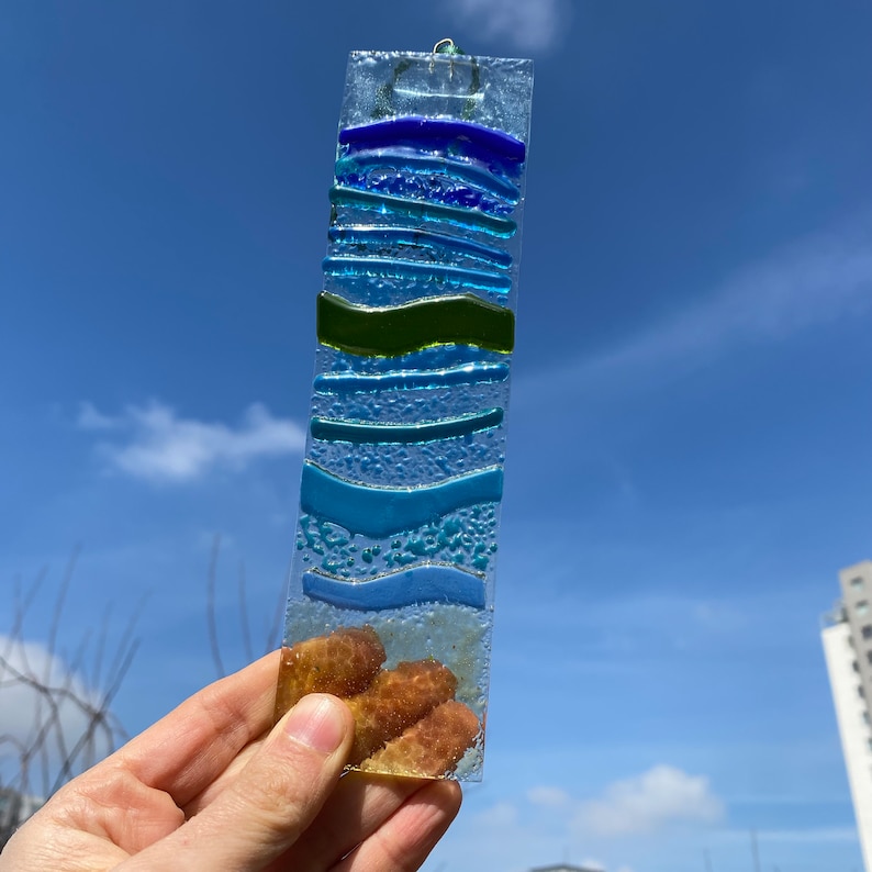 以下が含まれることがあります： 青、緑、茶色の海のテーマのデザインが施された、長方形のフュージングガラスのサンキャッチャー。ガラスは、青、緑、茶色の色合いなど、さまざまなテクスチャと色を持っています。吊り下げ用の小さな金属ループが上部にあります。