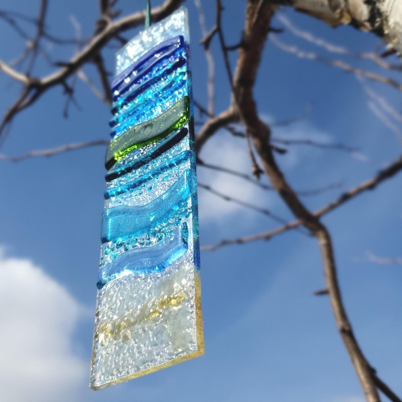 以下が含まれることがあります： 海岸をテーマにした長方形のフュージングガラスのアート作品。青、緑、白のガラスの層で構成され、海の波と砂浜を表現しています。緑色の紐で吊り下げられています。