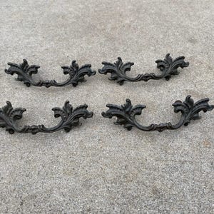 May include: Four antique bronze-colored drawer pulls with an ornate, floral design. Each pull features a curved handle with decorative leaf and scroll details, perfect for adding a vintage touch to furniture.