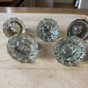 May include: Five antique glass door knobs with a faceted design. The knobs are clear glass with a slight green tint and have a brass base.