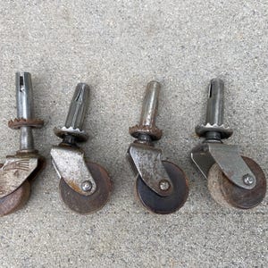 May include: Four vintage-style furniture casters featuring dark brown wooden wheels and weathered metallic silver-toned hardware. Each caster includes a vertical metal stem with a grooved top and a serrated circular collar above the wheel bracket.