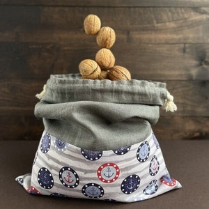 Può includere: Una borsa di tessuto grigio con coulisse e fodera a righe bianche e grigie. La borsa è decorata con ancore rosse, blu e bianche in cerchi. La borsa è piena di noci.