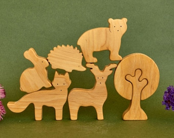 May include: Wooden animal figurines including a bear, deer, fox, rabbit, hedgehog, and tree. The figures are light brown and arranged on a green surface, with purple and pink flowers in the background.