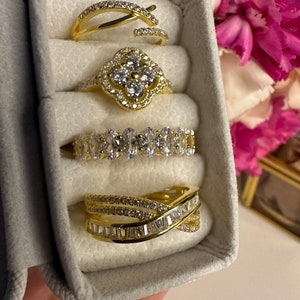 May include: Four gold rings with clear cubic zirconia stones in a white velvet ring box. The rings are stacked on top of each other.