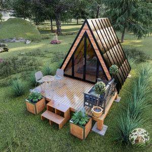 May include: A small, triangular wooden cabin with a black metal roof and large windows. The cabin is surrounded by green grass and trees. There are two chairs and a table on a wooden deck in front of the cabin.