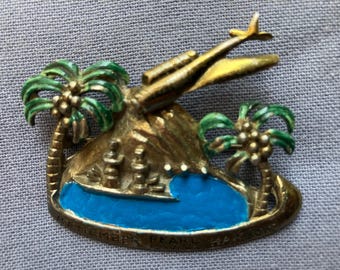 May include: A gold-toned metal brooch featuring a tropical island scene with two green palm trees, a blue enamel water section, a ship, and an airplane flying above. The base is engraved with the text REMEMBER PEARL HARBOR 12-7-41.