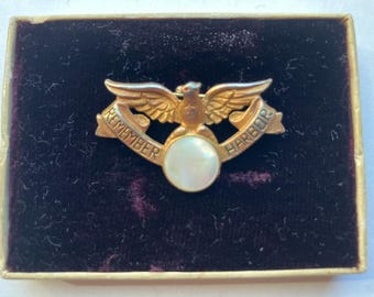 May include: A gold-toned pin with an eagle and the words "REMEMBER HARBOR". The pin has a white, round center and is displayed in a velvet-lined box.